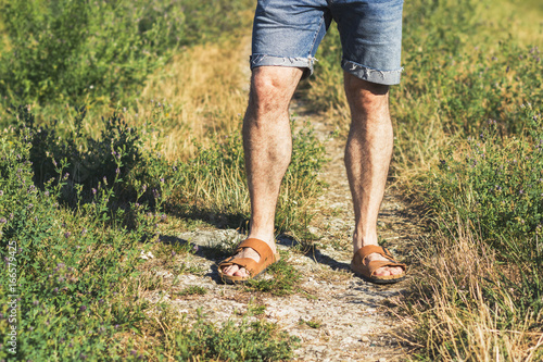 Male legs in brown leather sandals and blue jean shorts standing on a trail