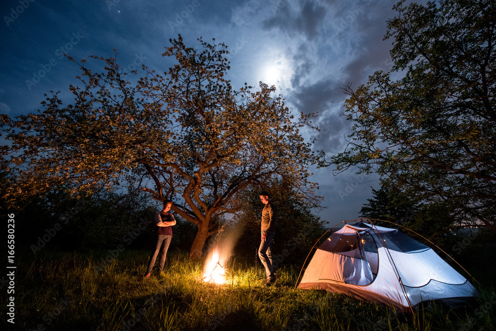 Romantic couple tourists standing at a campfire near tent under trees and night sky with the moon. Night camping - obrazy, fototapety, plakaty 