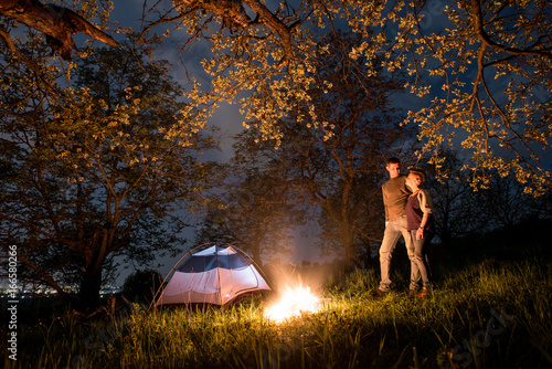 Romantic couple tourists standing at a campfire near tent, hugging and looking to each other under trees and night sky. Night camping