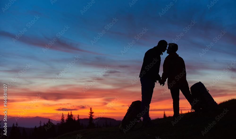 Silhouettes of a loving couple of hikers kissing on top of the mountain holding hands standing near their backpacks copyspace love anniversary achieving togetherness affection romantic hiking active.