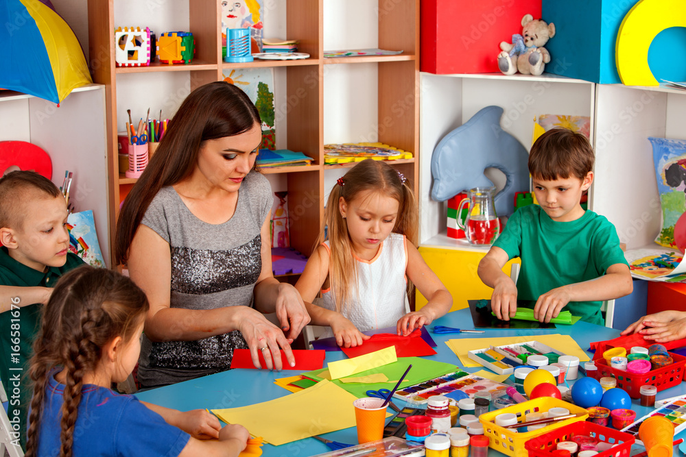 Fototapeta premium School children with scissors in kids hands cutting paper with teacher class room. Group of little ones in an orphanage. Children's project in kindergarten. Kids play into team.