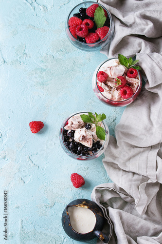 Traditional summer dessert Eton Mess. Broken meringue with whipped cream, berry jam, fresh blueberries and raspberries in two glasses, decorated with mint leaves over light blue concrete background.