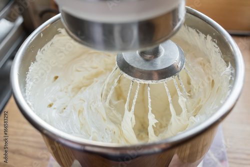 Closeup of electric mixer with whipped smooth dough for cake. Batter being whipped. Mixing white dough in bowl with motor mixer, baking cake