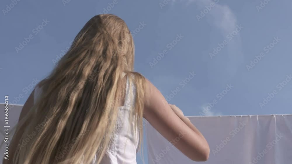 Rear view of young girl hanging up wet clean laundry on a clothesline against blue sky. Girl's long hair and white bed sheets on washing line blowing in the wind. Low angle view. Rear view of young girl hanging up wet clean laundry on a clothesline against blue sky. Girl's long hair and white bed sheets on washing line blowing in the wind. Low angle view.