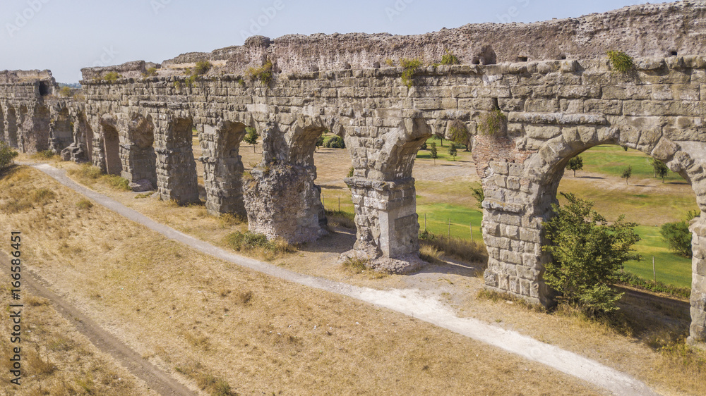Vista aerea laterale dei resti di un'acquedotto romano situato nel parco degli Acquedotti a Roma, vicino la fermata metro Cinecittà. In primo piano dei campi di grano.