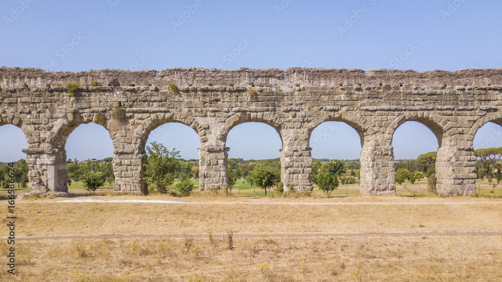 Vista aerea frontale dei resti di un'acquedotto romano situato nel parco degli Acquedotti a Roma, vicino la fermata metro Cinecittà. In primo piano dei campi di grano.