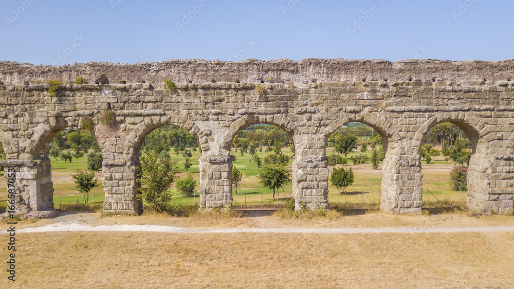 Vista aerea frontale dei resti di un'acquedotto romano situato nel parco degli Acquedotti a Roma, vicino la fermata metro Cinecittà. In primo piano dei campi di grano.