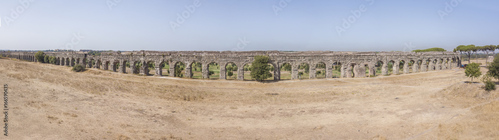 Vista aerea frontale e panoramica dei resti di un'acquedotto romano situato nel parco degli Acquedotti a Roma, vicino la fermata metro Cinecittà. In primo piano dei campi di grano.