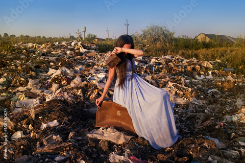 A girl with an empty wallet sits on a suitcase among the garbage dump and cries