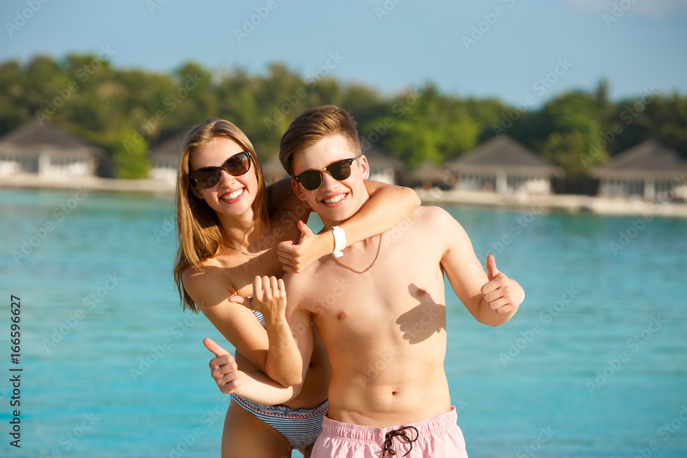 Happy young couple have fun and relax on the beach. Man and woman show thumbs up enjoying vacation near ocean. Bungalows of spa resort, palms, and blue lagoon on background. Family travel concept Happy young couple have fun and relax on the beach. Man and woman show thumbs up enjoying vacation near ocean. Bungalows of spa resort, palms, and blue lagoon on background. Family travel concept
