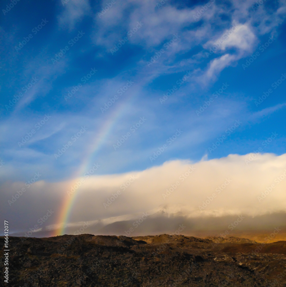 Beautiful landscape view of Real Natural Colorful Rainbow over the ancient icelandic mountain ranges hidden in misty clouds and blue sky background, Olafsvik, Iceland.