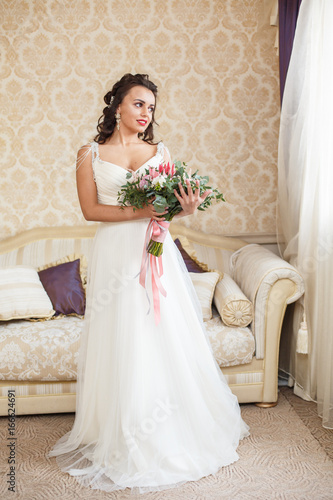Beautiful young Bride with dark hairs in a bedroom. Classic white wedding dress. Half-length portrait