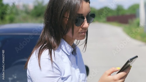 Moving portrait of woman at summer time. Yoyng strict female in rounded black sunglasses stay near her black car at sunny day. photo