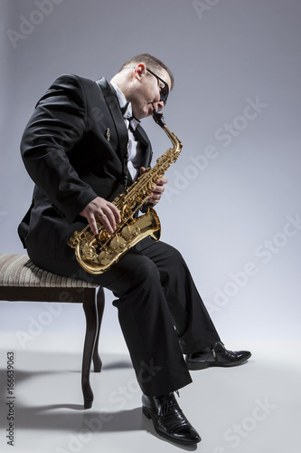 Music Concepts. Portrait of Mature Relaxed and Thoughful Caucasian Saxophone Player in Sunglasses Playing the Saxophone While Sitting on Chair in Studio Environment.