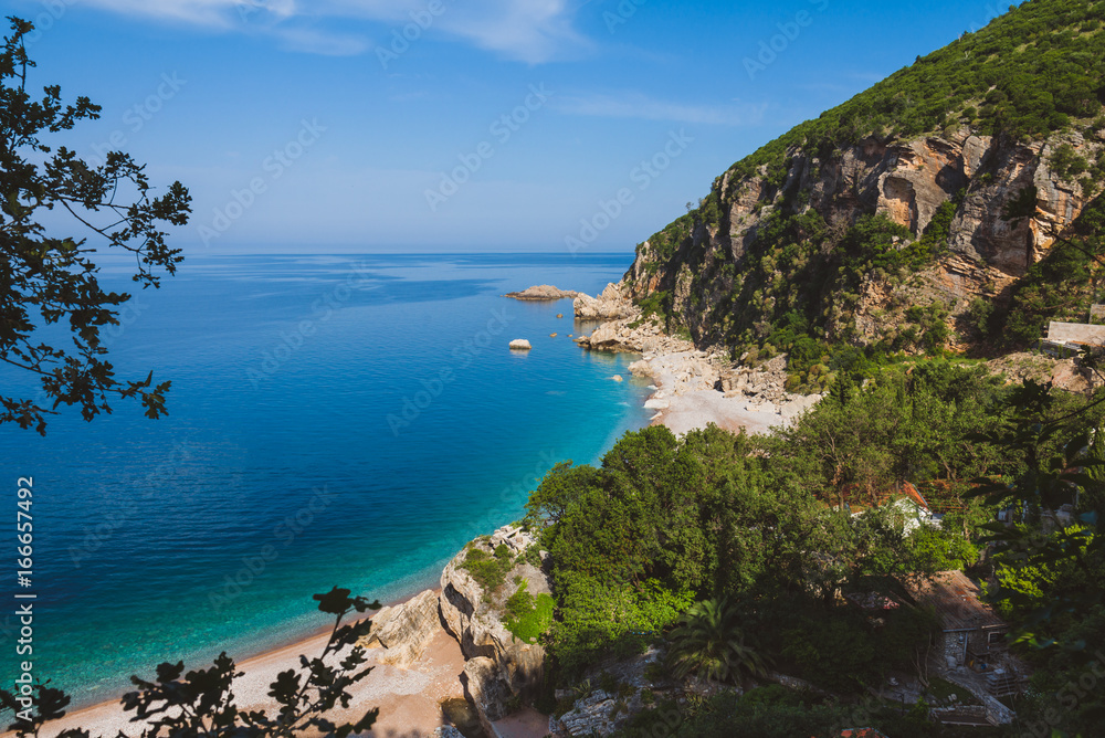 Adriatic sea coast by sunny day summer landscape. Pebble beach, house roofs and green tree brunches under blue sky near Perazica Do village on the way to Petrovac, Montenegro.