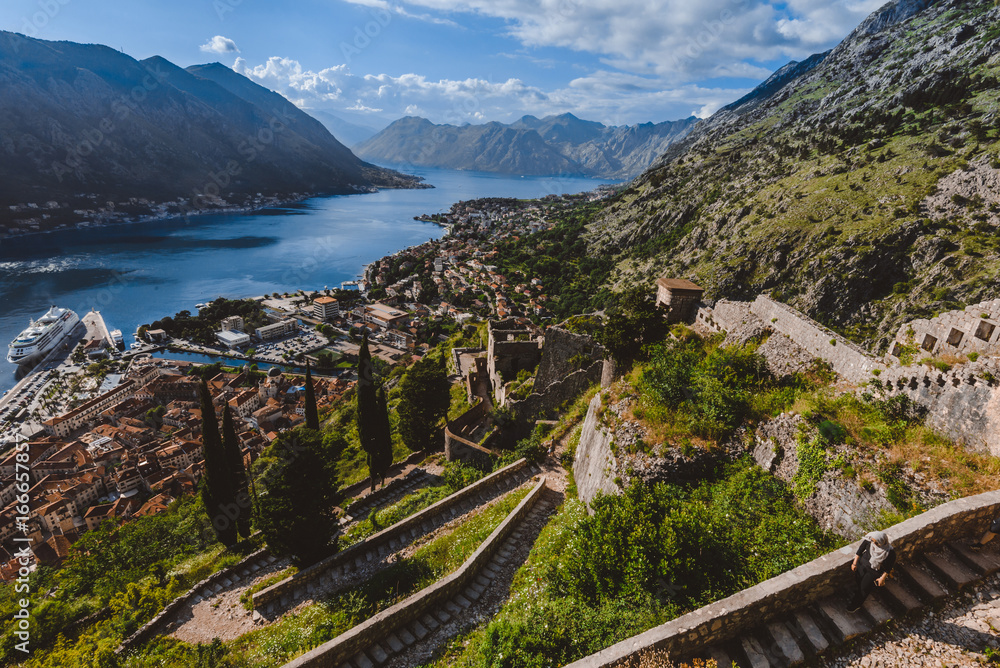 Fototapeta premium Kotor fortress walls and stone staircase wide angle view with Kotor Bay, Old Town rooftops and cloudy mountains on background. Boka Kotorska, Montenegro.