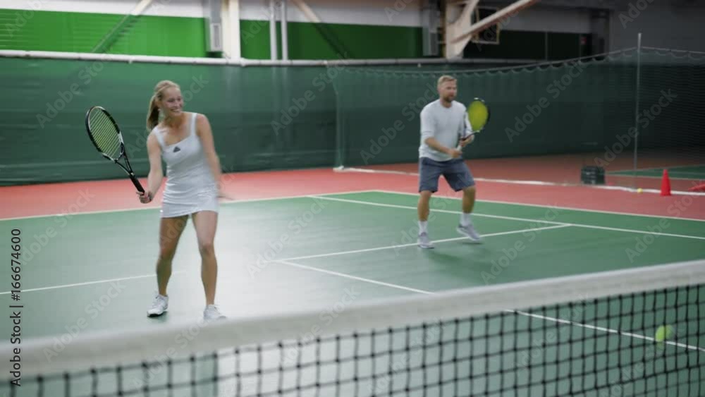 Image of happy couple playing tennis at indoor court. Young people dressed in sport outfit are spending time together training in recreation area. Man and woman holding rackets and hitting balls. Image of happy couple playing tennis at indoor court. Young people dressed in sport outfit are spending time together training in recreation area. Man and woman holding rackets and hitting balls.
