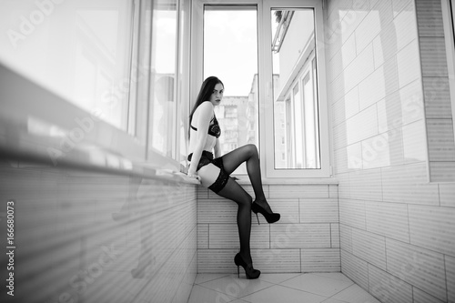 Portrait of an awesome young woman sitting on the windowsill in lingerie  stockings and high heels. Black and white photo.