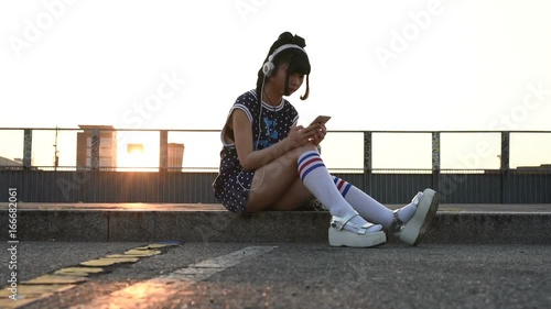 Young beautiful asian nonconformist woman listening music with headphones and smart phone hand hold outdoor in city back light, tapping the touchscreen - music, technology, social network concept