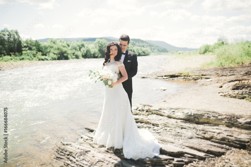Beautifull wedding couple kissing and embracing near the shore of a mountain river with stones Beautifull wedding couple kissing and embracing near the shore of a mountain river with stones