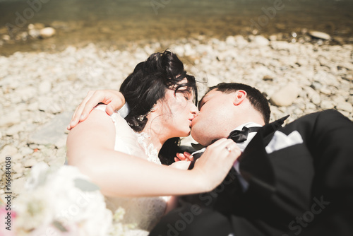 Close up portrait of couple against river and green trees. Beautiful young woman kissing handsome man outdoors