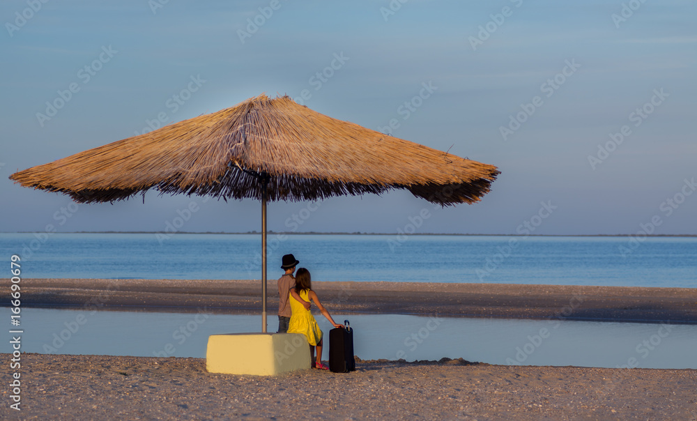 A boy in a hat and a girl on the beach with a suitcase under a straw umbrella look into the distance. Traveling of children in the summer holidays