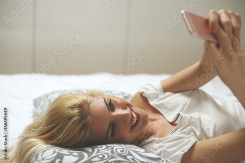 Young woman lying in bed and typing on smart phone. Cheerful woman using mobility.