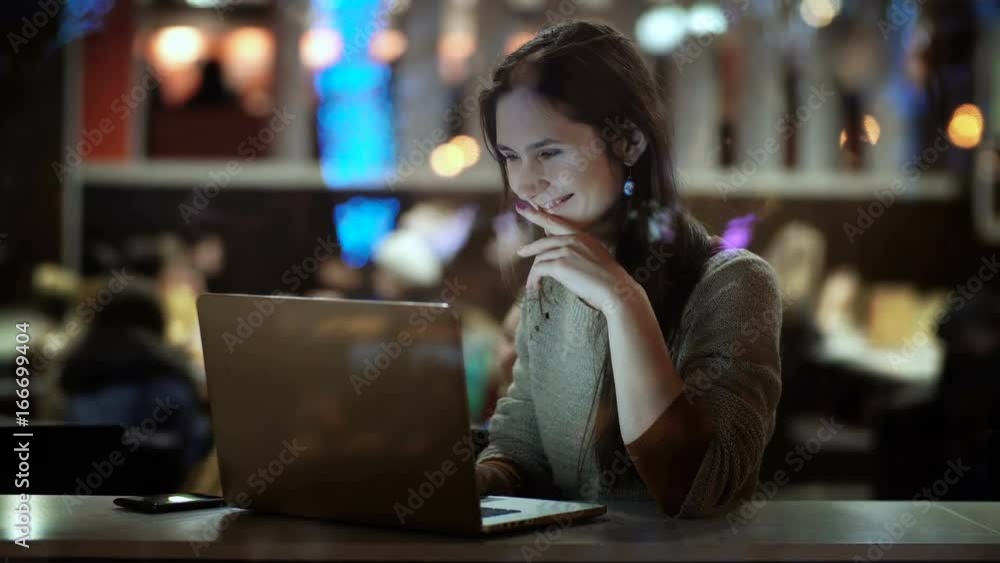 View inside the window. Young happy woman sitting in the evening at cafe and using the laptop computer, smiling. View inside the window. Young happy woman sitting in the evening at cafe and using the laptop computer, smiling.