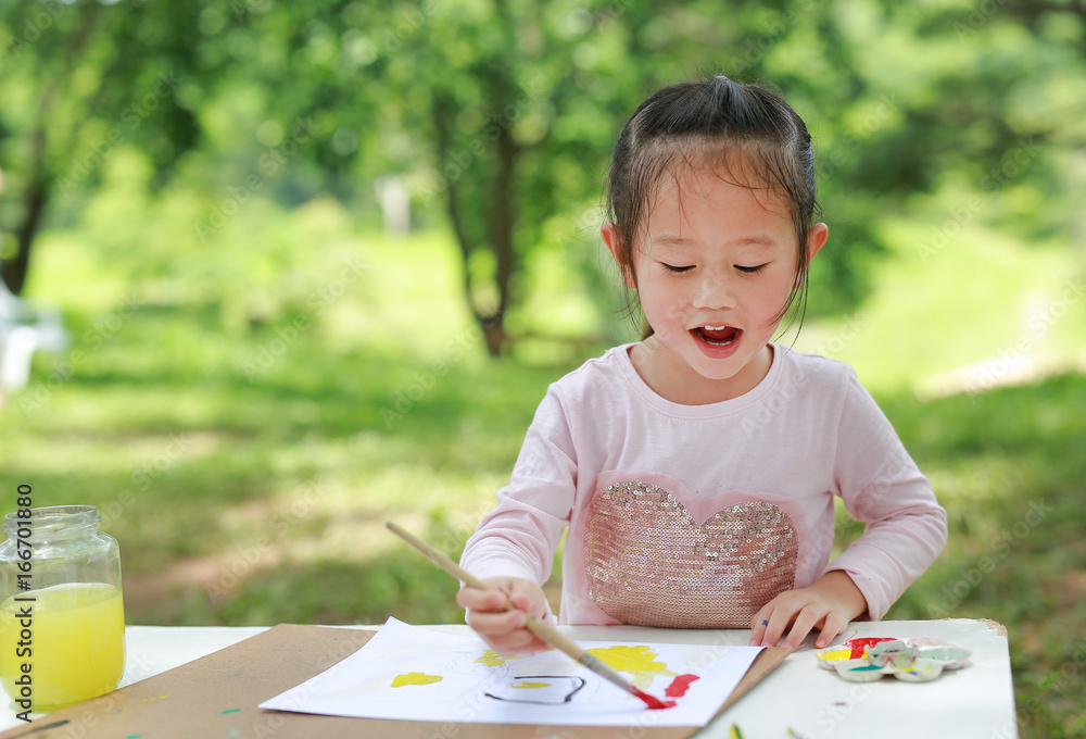 Happy asian child girl sitting at a table in a summer garden painting with paintbrush, Education art concept. Happy asian child girl sitting at a table in a summer garden painting with paintbrush, Education art concept.