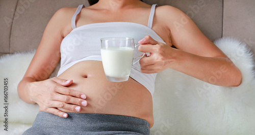 Young pregnant woman with glass of milk sitting on sofa in the room.