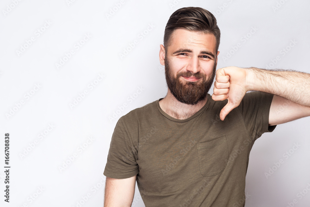Portrait of unsatisfied bearded man with thumbs down and dark green t shirt against light gray background. studio shot. . Portrait of unsatisfied bearded man with thumbs down and dark green t shirt against light gray background. studio shot. .