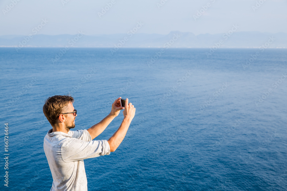 Handsome young man wearing sunglasses taking a photo of the horizon at sea Handsome young man wearing sunglasses taking a photo of the horizon at sea