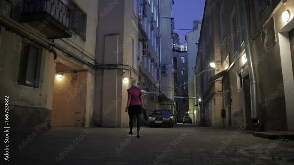 shortcut blondie woman goes out from inner yand to the street at dark night. There are electric lights on buildings. She wears black dress, pink jacket, clutch bag, Saint Peterburg, Russia shortcut blondie woman goes out from inner yand to the street at dark night. There are electric lights on buildings. She wears black dress, pink jacket, clutch bag, Saint Peterburg, Russia