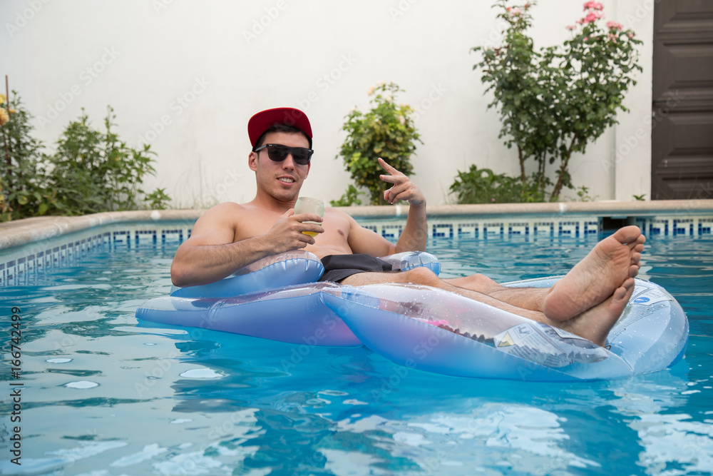 Young strong man drinking beer on an air bed in the pool. 