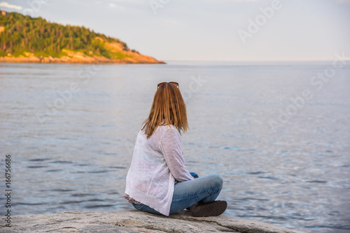woman sitting on rocky shore of Tadoussac, Quebec looking for whales at sunset