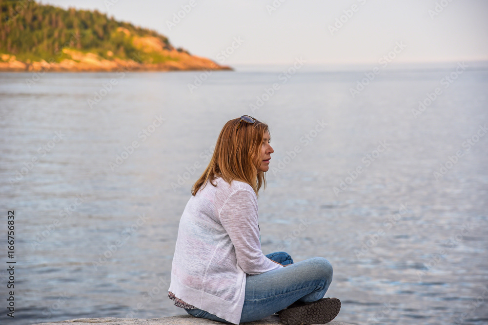 Woman sitting on rocky shoreline in Tadoussac, Quebec looking for whales