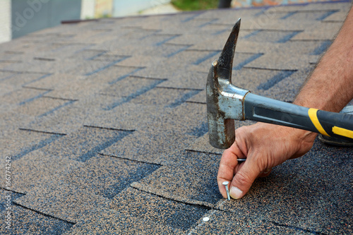 Worker hands installing bitumen roof shingles. Worker Hammer in Nails on the Roof. Roofer is hammering a Nail in the Roof Shingles. Unfinished roof.