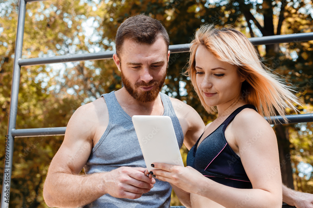 Young athletic girl and bearded man browsing the internet on tablet PC while doing fitness exercises in park at autumn day. Young athletic girl and bearded man browsing the internet on tablet PC while doing fitness exercises in park at autumn day.
