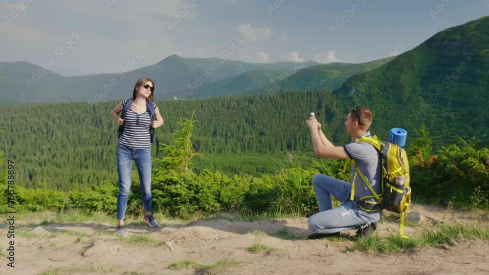 A young couple is photographed in a picturesque mset on the background of the mountains covered with forest. The guy takes pictures of his girlfriend A young couple is photographed in a picturesque mset on the background of the mountains covered with forest. The guy takes pictures of his girlfriend