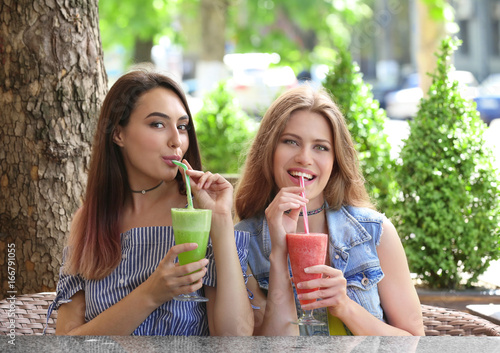 Two positive young women enjoying fresh smoothie in cafe