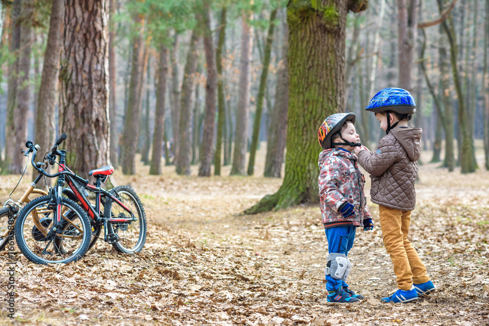Obraz premium Two brothers preparing for bicycle riding in spring or autumn forest park. Older kid helping sibling to wear helmet. Safety and protection concept. Happy boys best friends having good time together.