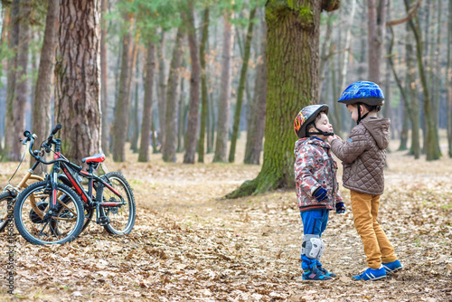 Two brothers preparing for bicycle riding in spring or autumn forest park. Older kid helping sibling to wear helmet. Safety and protection concept. Happy boys best friends having good time together.