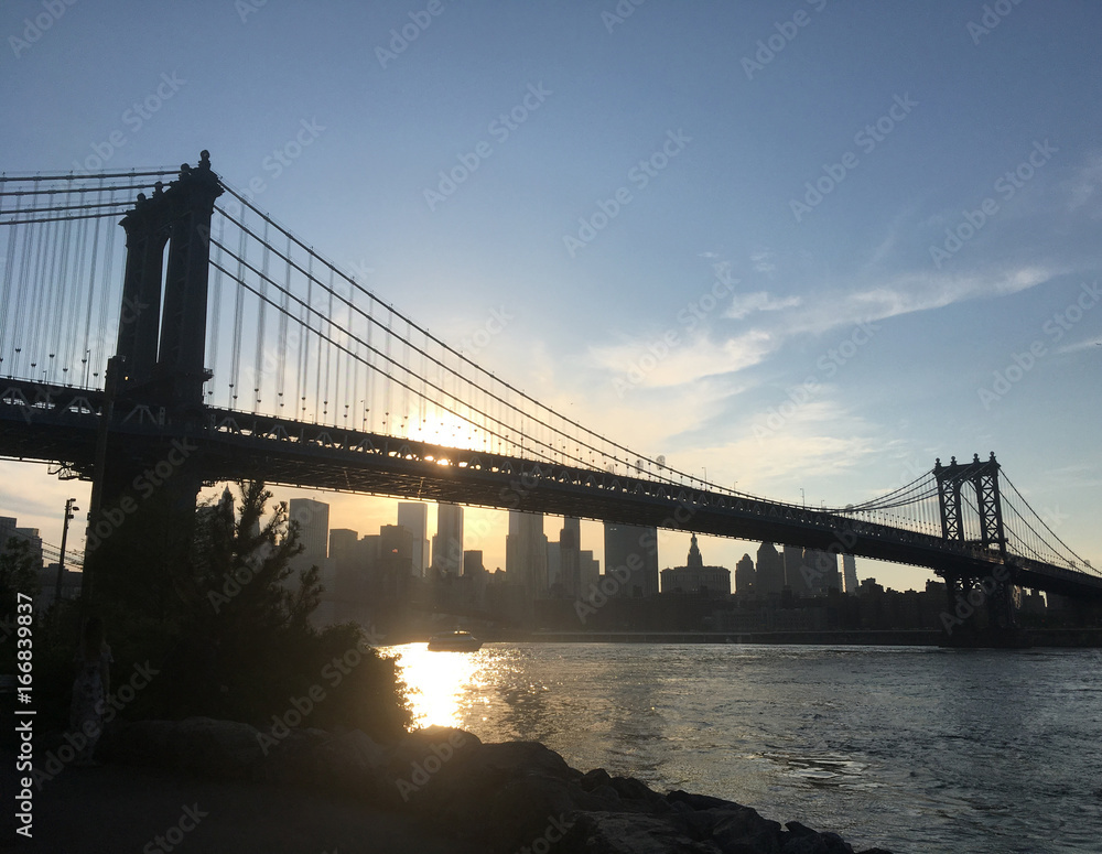 Sunset sets over the Manhattan Bridge and the East River, view from Brooklyn shore. Manhattan Bridge in silhouette against setting sun, New York Financial district and skyscrapers at sunset.