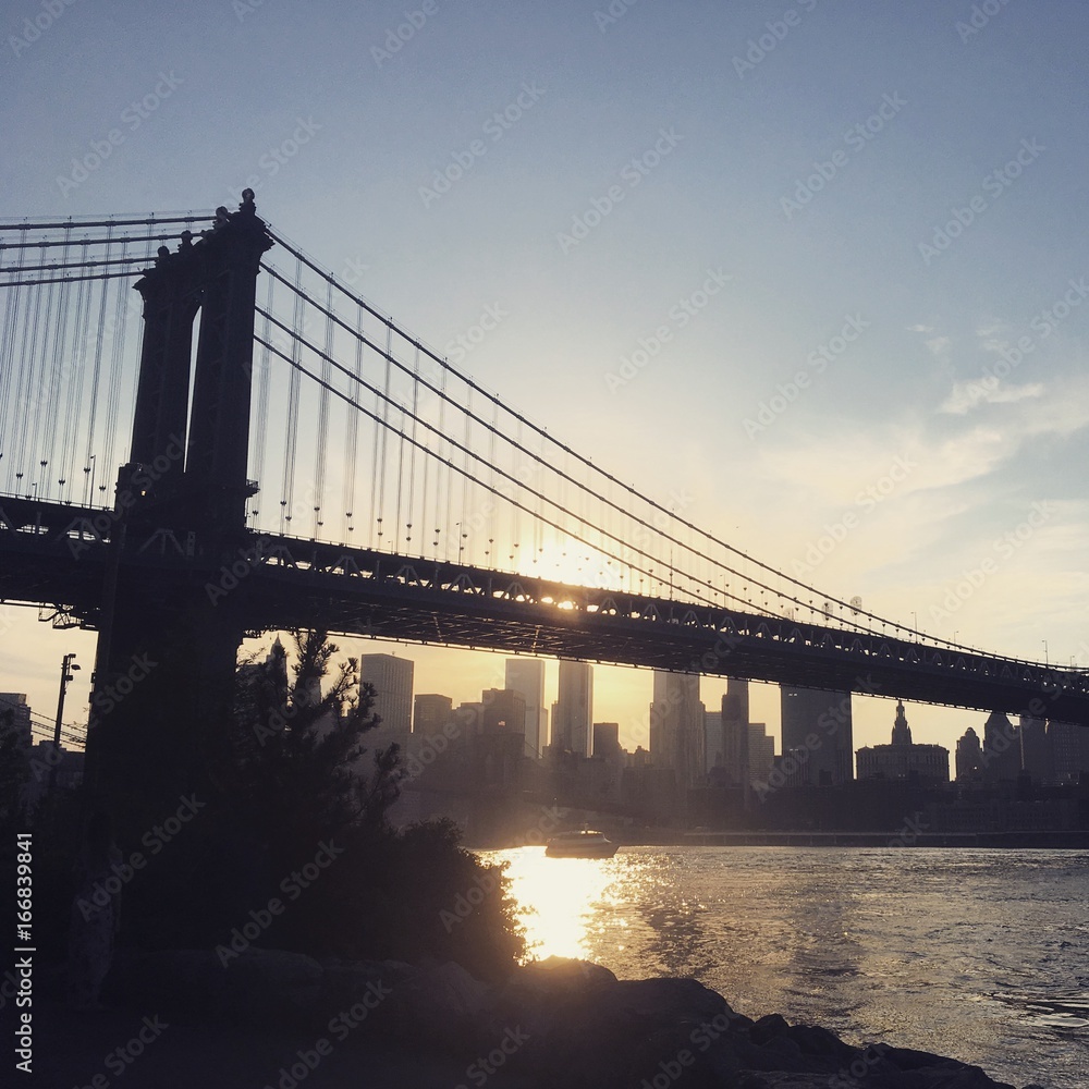 Obraz premium View of the Manhattan Bridge at sunset from Brooklyn shoreline. Looking towards New York Financial District at sunset, Manhattan Bridge spans the East River as the sun sets.