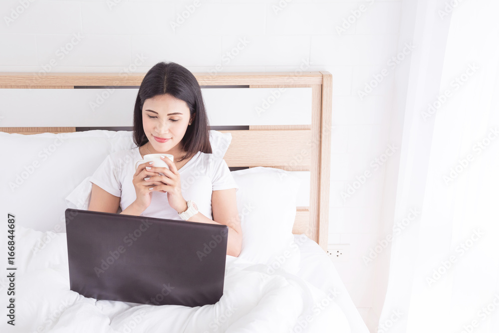 Beautiful asian woman sitting on bed ,drinking coffee and using laptop computer Beautiful asian woman sitting on bed ,drinking coffee and using laptop computer