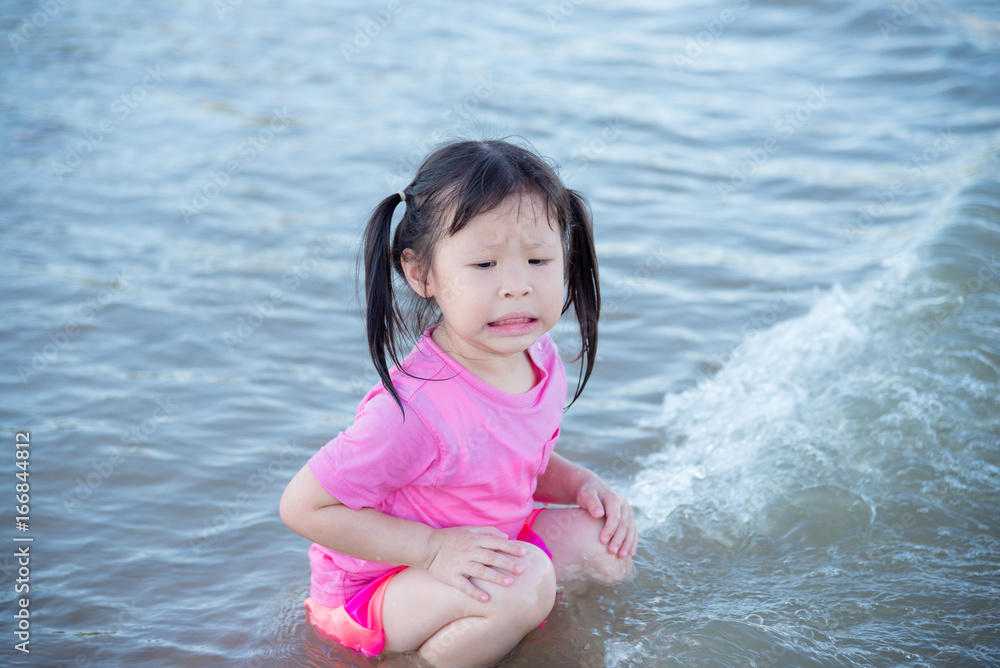 Little Asian girl shocked after being hit by the sea waves while sitting by the seashore