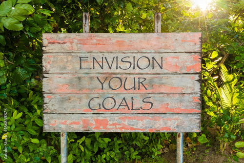 Envision Your Goals motivational quote written on old vintage board sign in the forrest, with sun rays in background.