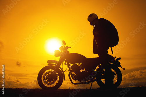 Silhouette of biker man  with his motorbike he shoulder backpack. beside the natural lake and beautiful  enjoying freedom and active lifestyle  having fun on a bikers tour.sunset background and sky.