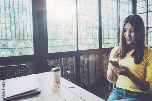 Happy Asian woman chatting on her mobile phone while relaxing in cafe during free time, Charming and beautiful hipster girl with smile reading good news on smart phone during rest in a coffee shop.