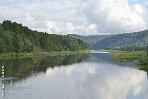 Summer river landscape. A small calm river. The hilly banks were overgrown with forest. Along the left bank the electric wires are stretched.